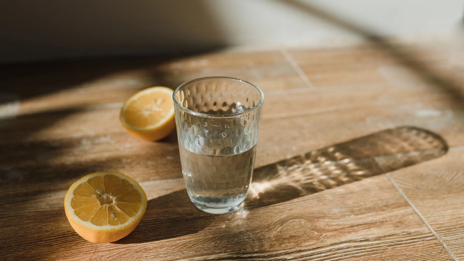 A glass of water on a wooden table in low morning sunlight, beside two lemon halves